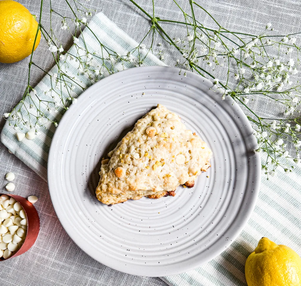 A sourdough scone with white chocolate and lemon on a plate next to white chocolate chips, a lemon and flowers.