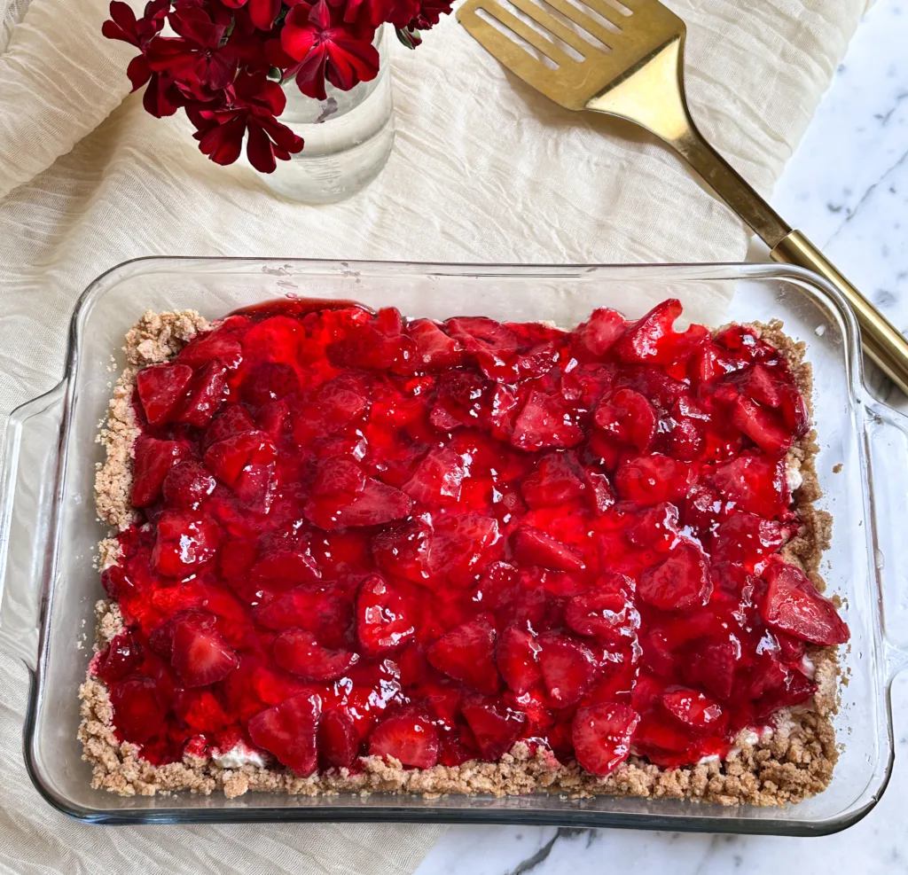 A pan of no-bake strawberry cheesecake next to a metal spatula and some red flowers.