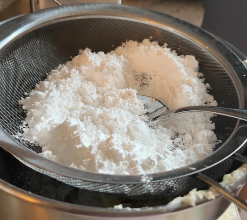 A fine mesh strainer with powdered sugar on top of a stand mixer bowl.