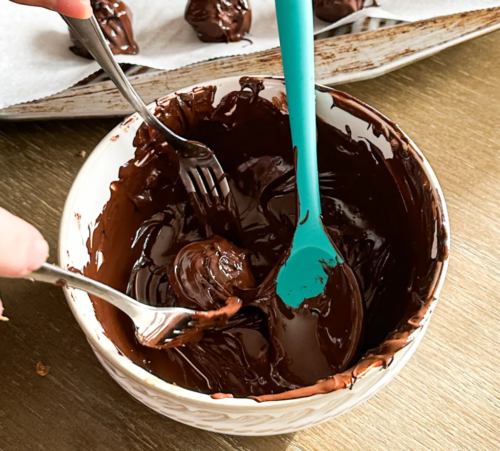 A bowl with melted chocolate and two forks dipping a date ball into the melted chocolate.