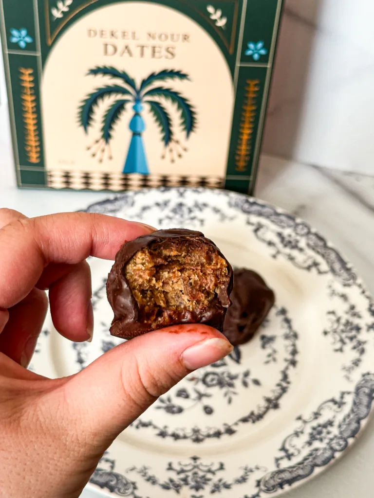 A chocolate date ball with a bite taken out of it and a plate with a date box in the background.