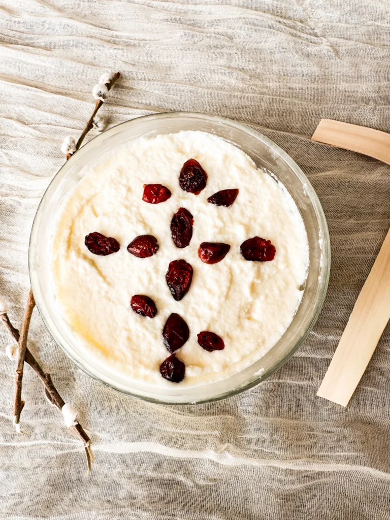 A bowl of cheese pascha, decorated with crasins and sitting next to a cross and a willow branch.