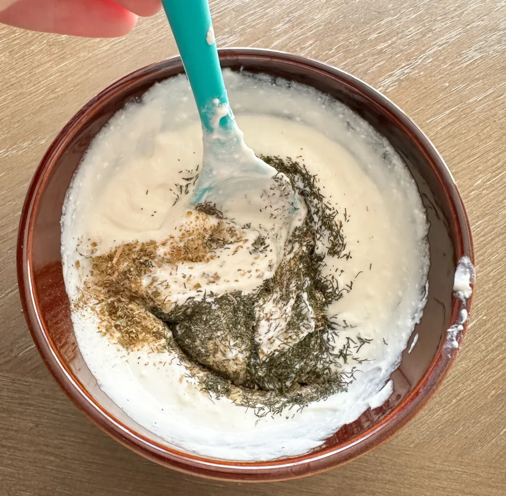 A spoon mixing together sour cream, herbs, garlic salt, and pepper together in a bowl.