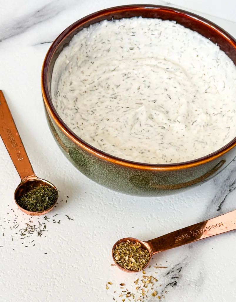A bowl of sour cream dip next to 2 measuring spoons with herbs in them.
