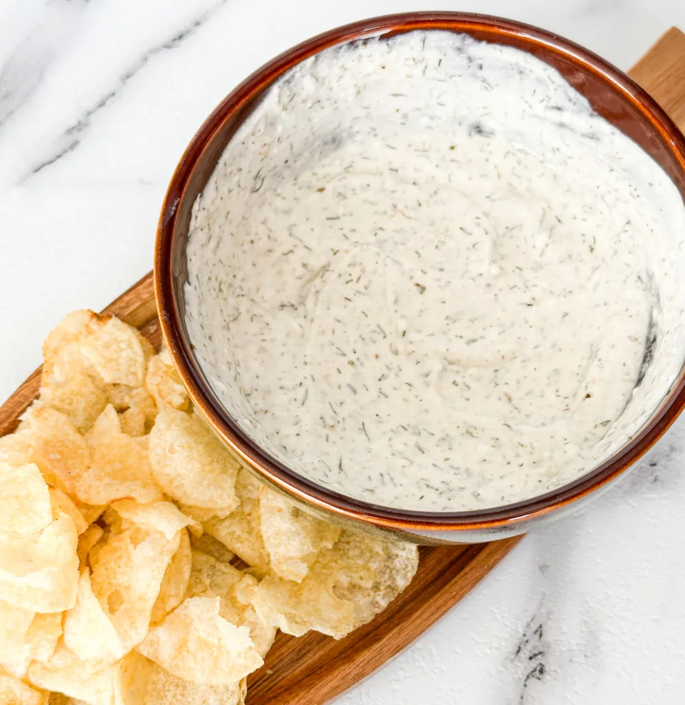 A bowl of sour cream dip on a wooden tray with potato chips.