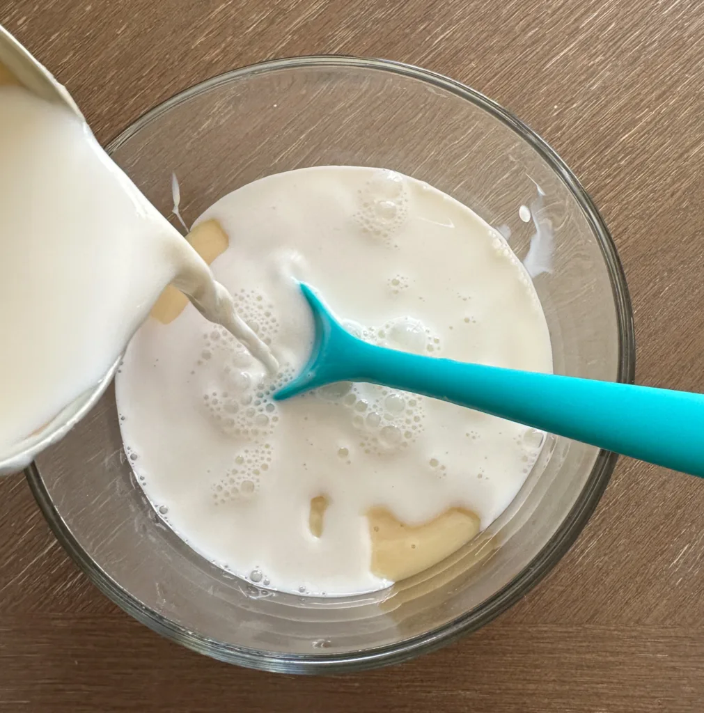 A cup of milk being poured into a bowl with sweetened condensed milk in it.