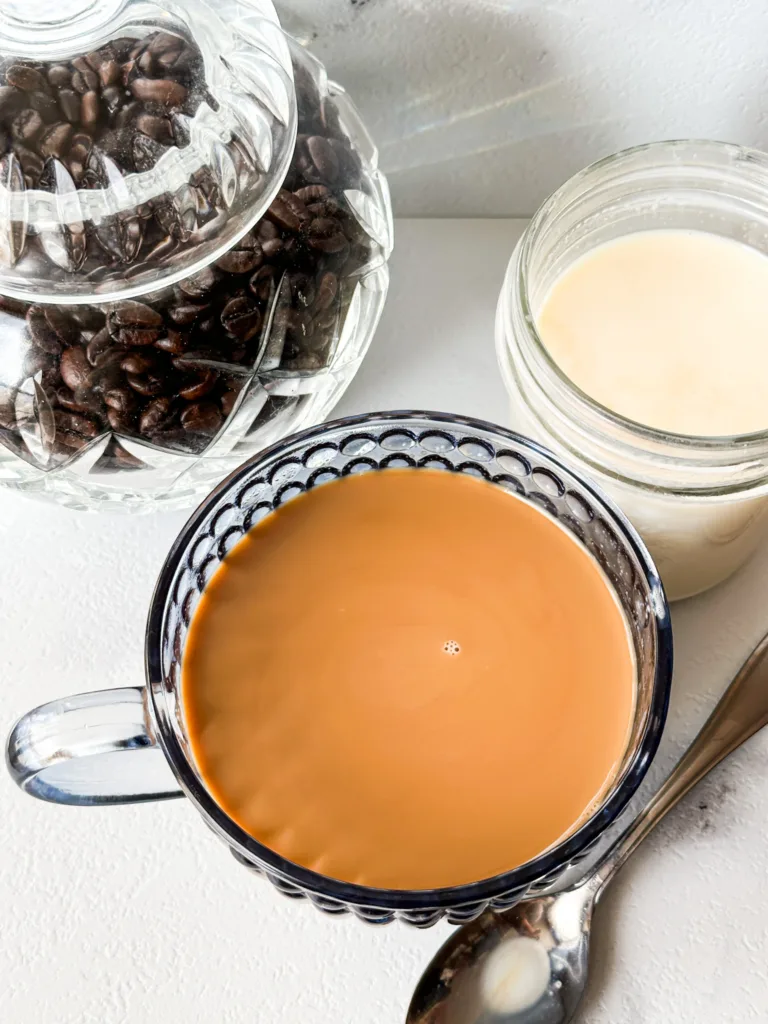 A cup of coffee next to a container of coffee beans and a jar of creamer.