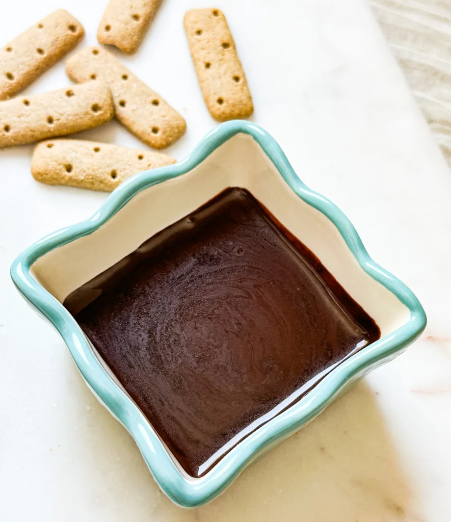 A chocolate sauce recipe in a square bowl next to some graham crackers.