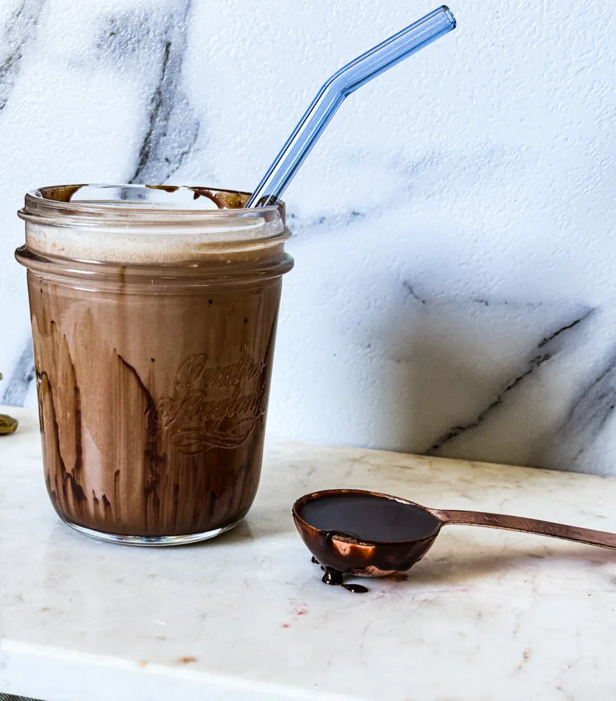 My chocolate sauce recipe in a table spoon next to a glass of milk with chocolate sauce in it.