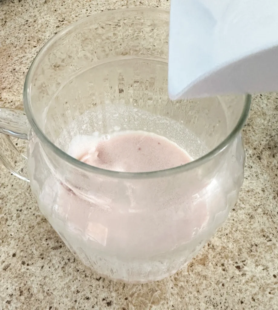 A glass cup with strawberry syrup and milk with ice being added to it.