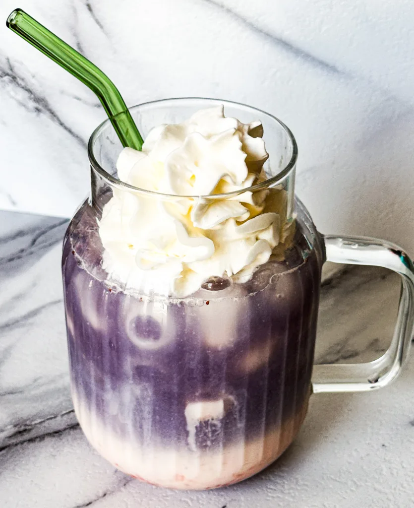 Butterfly pea tea latte in a glass cup with a straw and whipped cream.