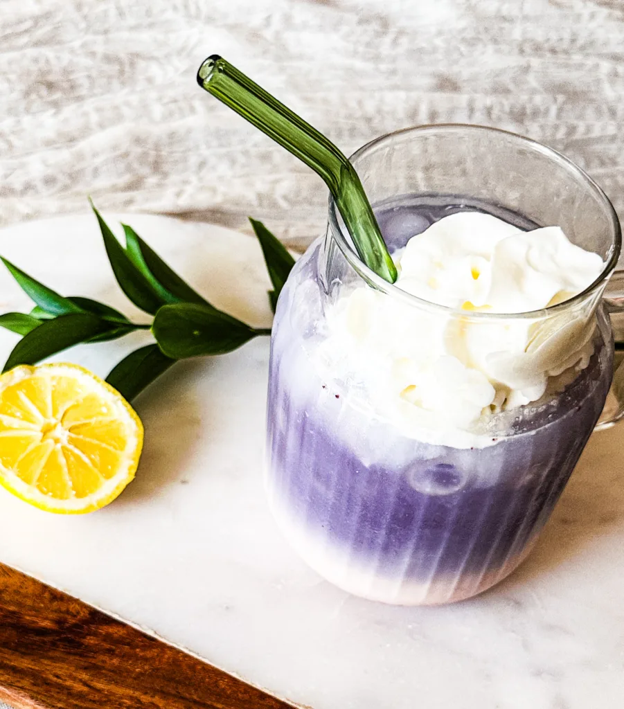 Butterfly pea tea latte on a marble cutting board with some greenery and half of a lemon.