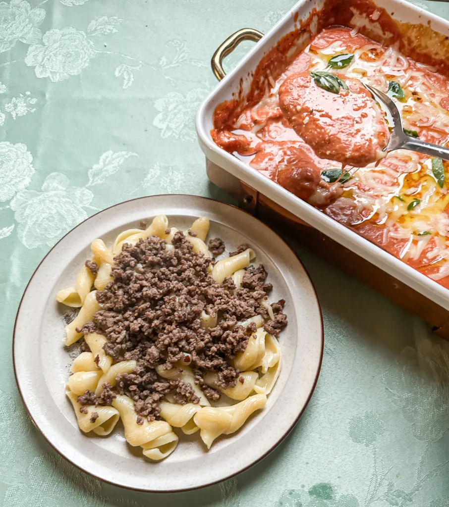 A pan of cream cheese pasta sauce with a spoon hovering over and a plate of ground beef and noodles next to it.