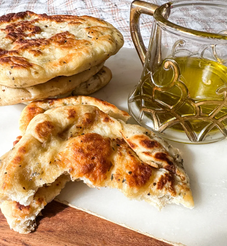 Pieces of homemade naan recipe on a cutting board next to a small bowl of olive oil.