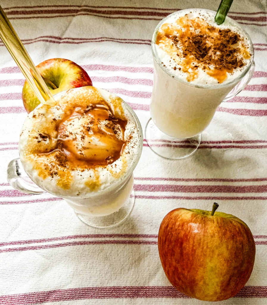 Two apple dirty soda floats in glasses with cinnamon and caramel on top, next to some apples.