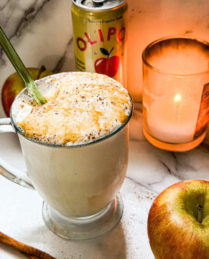 An apple dirty soda float in glass with a straw next to a Crisp Apple Olipop, a candle, and some apples.